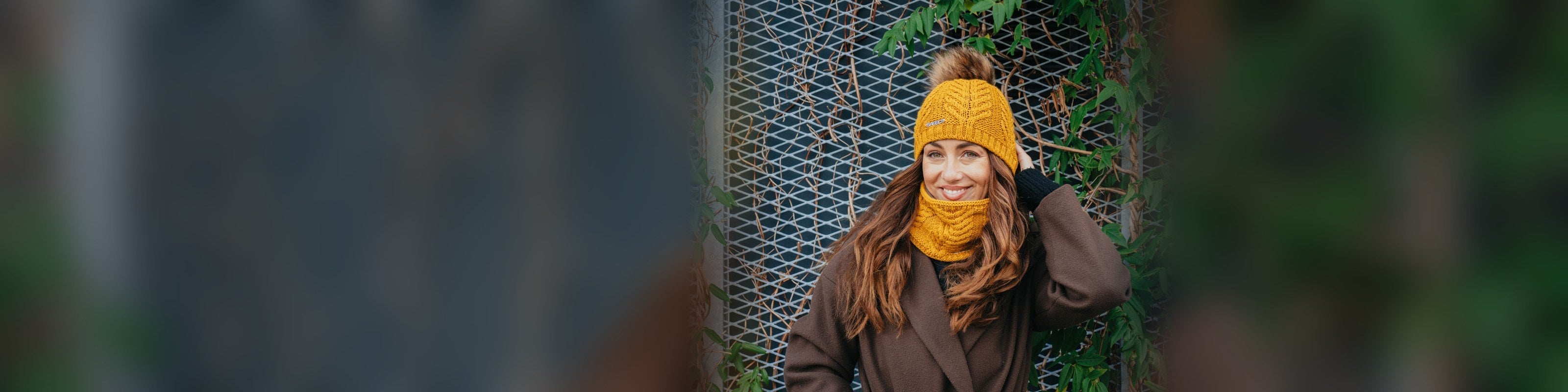 Woman smiling in a mustard SABBOT Emma cable-knit bobble hat with a faux fur pom and matching snood