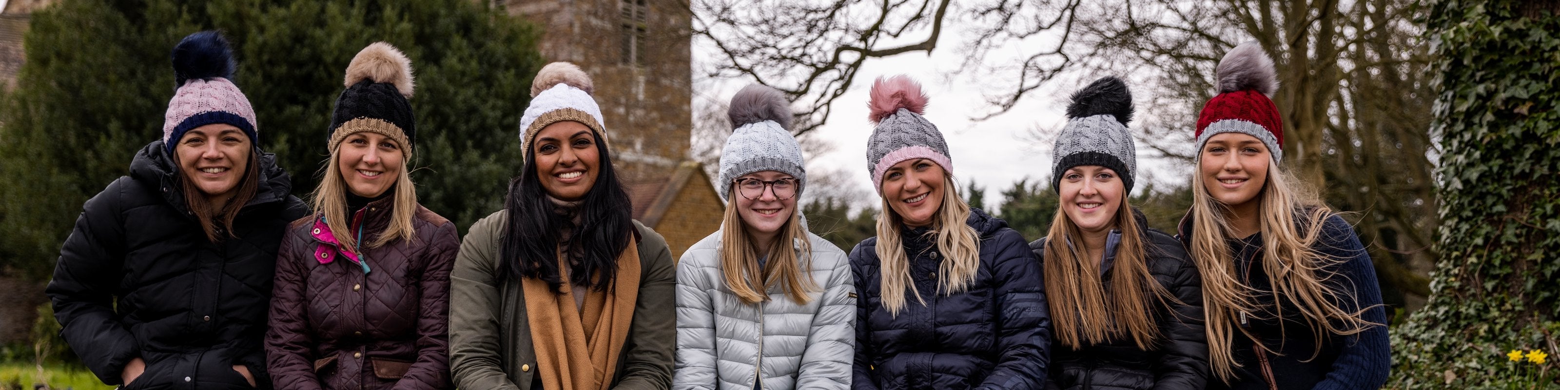 Seven women sitting on a churchyard wall wearing SABBOT bobble hats with faux fur poms