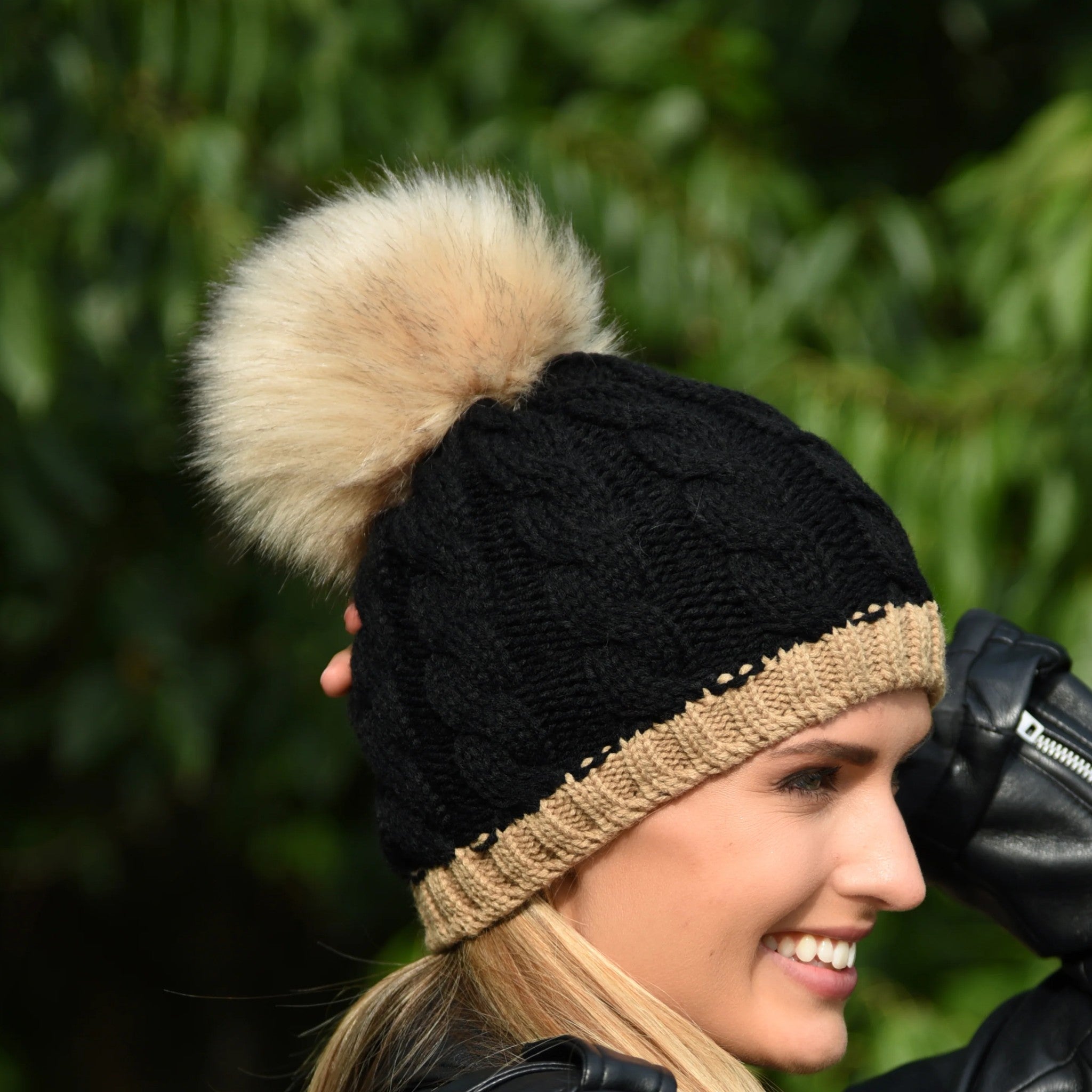 A woman in a black leather jacket wearing the Andrea chunky knit bobble hat in Black/Camel with faux fur pom pom, by Sabbot