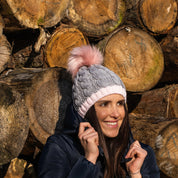 A woman in front of stacked logs wearing the Andrea chunky knit bobble hat with pink faux fur pom pom, by Sabbot