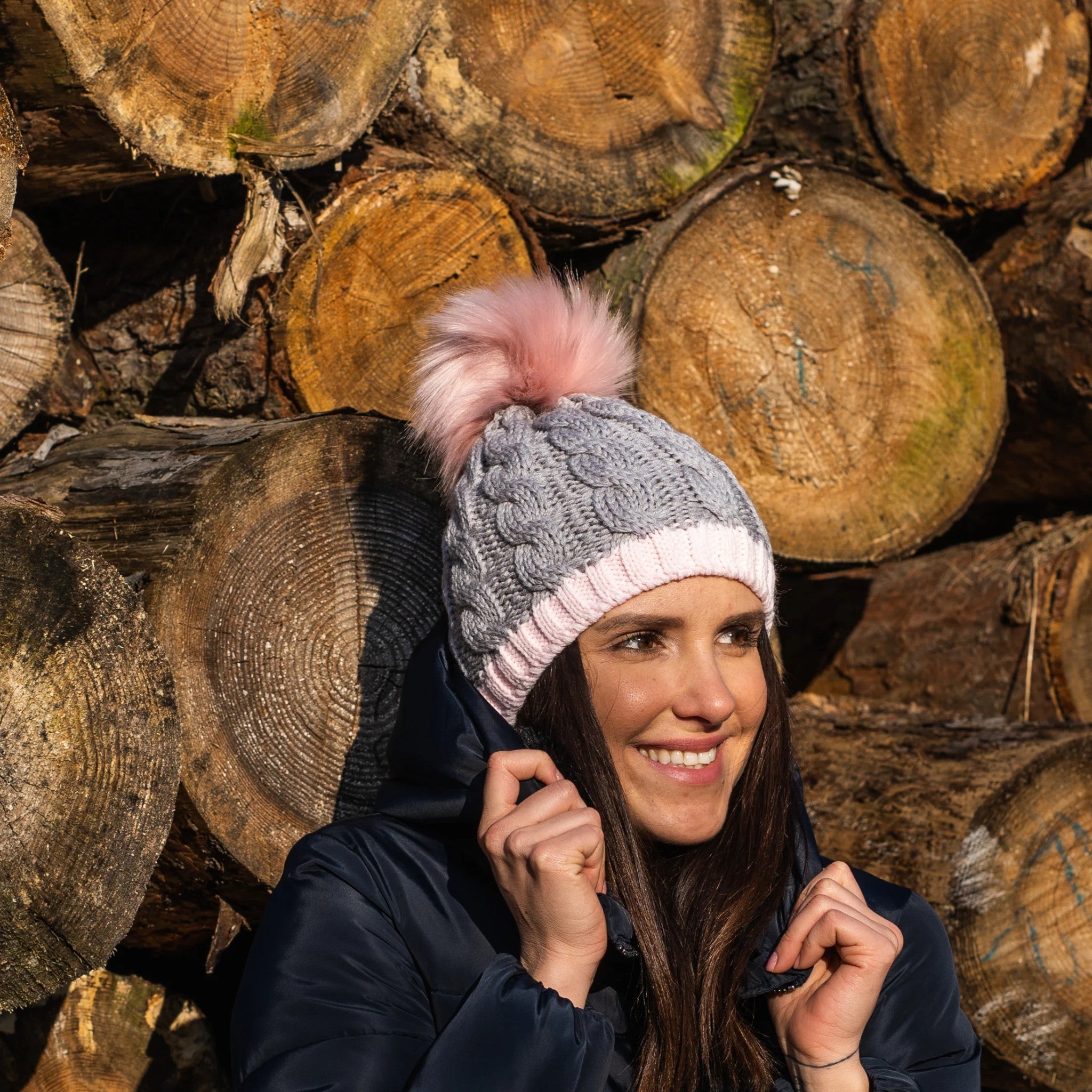 A woman in front of stacked logs wearing the Andrea chunky knit bobble hat with pink faux fur pom pom, by Sabbot