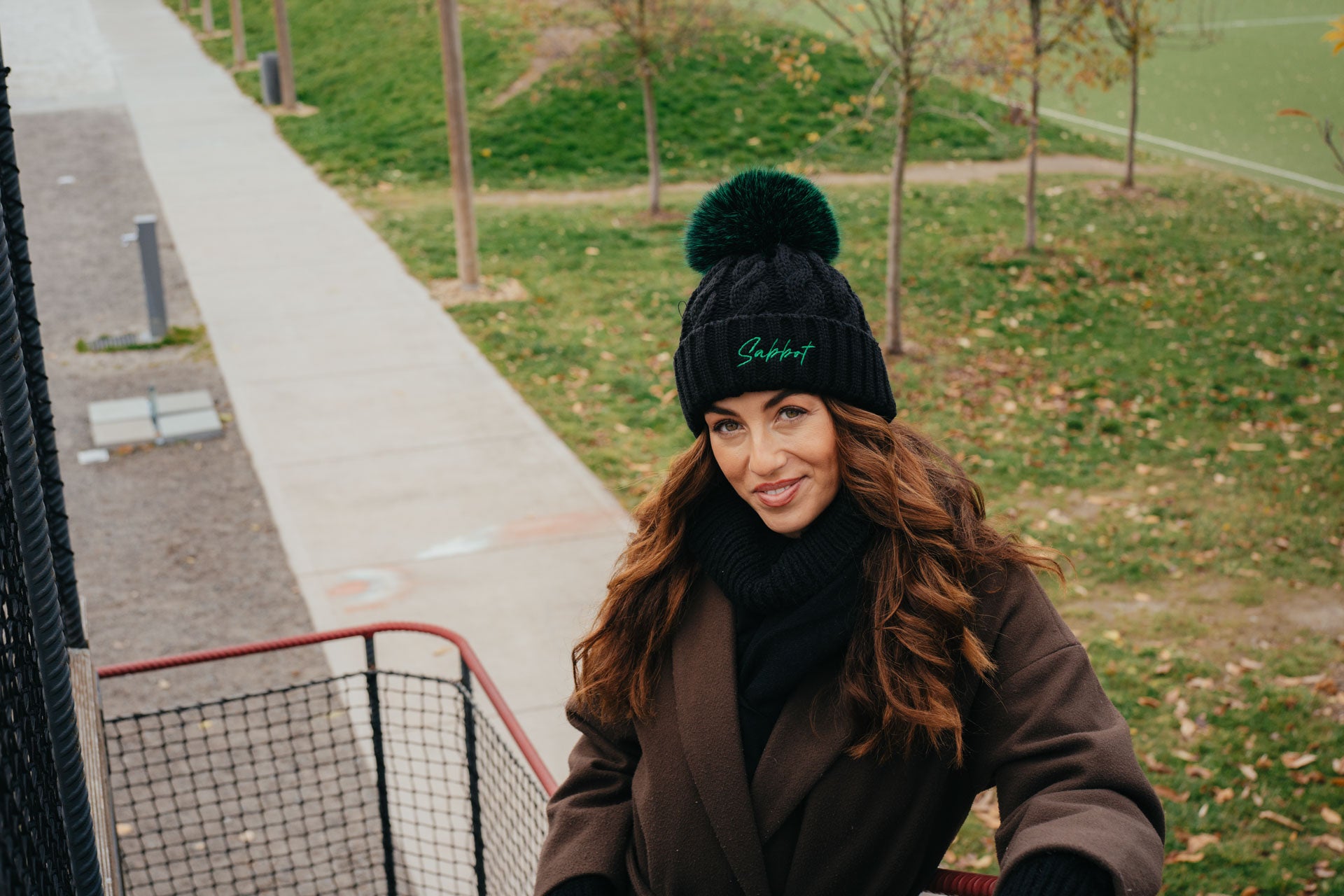 Sabbot Headwear Woman wearing a black beanie with a green faux fur pom-pom and brown coat in an outdoor setting