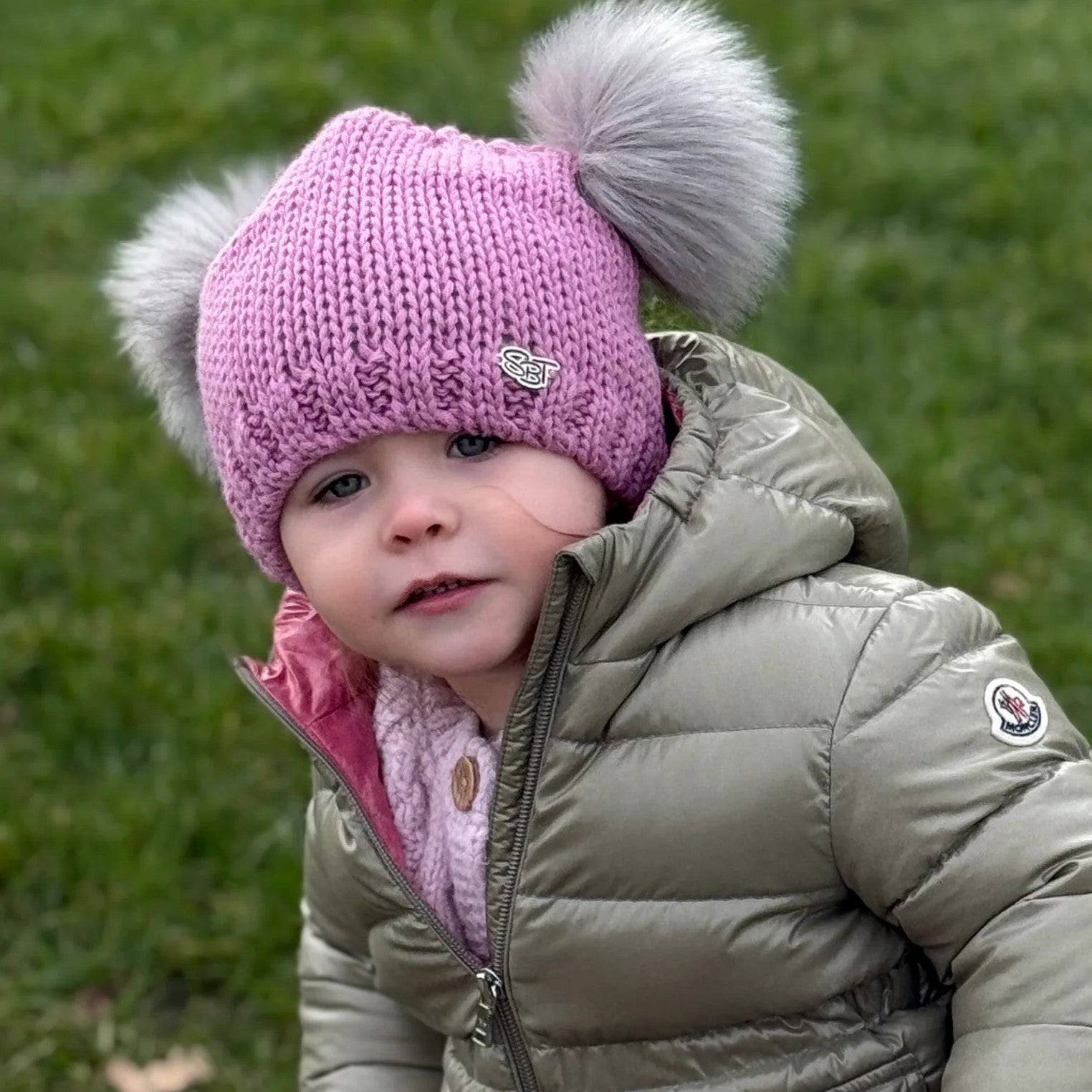 Toddler in a puffer jacket wearing the pink SABBOT Frankie children's bobble hat with two grey faux fur pom-poms