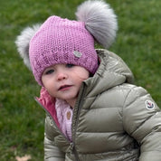 Toddler in a puffer jacket wearing the pink SABBOT Frankie children's bobble hat with two grey faux fur pom-poms