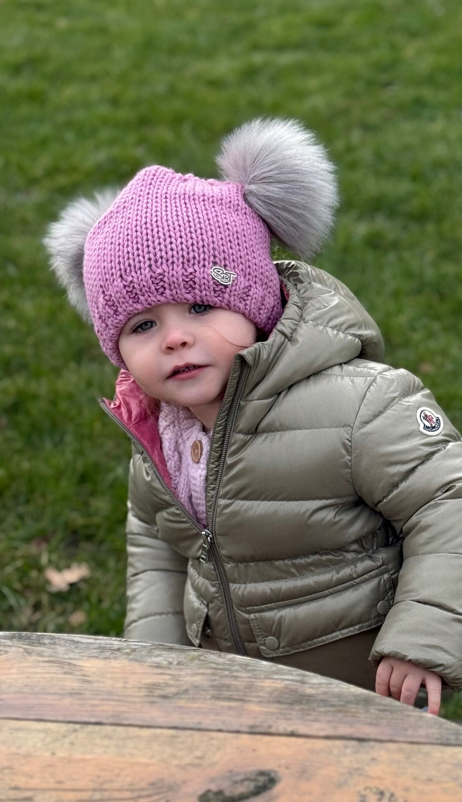 Toddler in a puffer jacket wearing the pink SABBOT Frankie children's bobble hat with two grey faux fur pom-poms