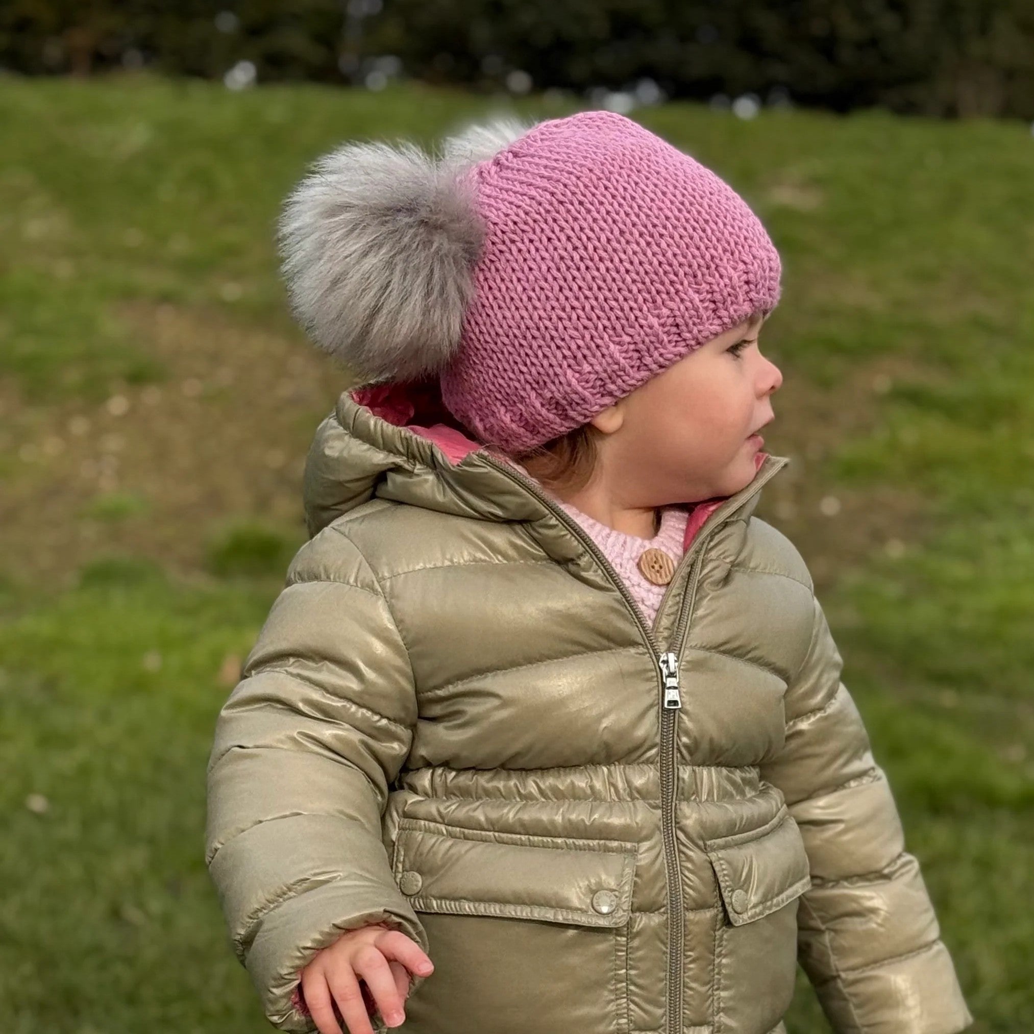 Toddler on a grassy hillside wearing a pink SABBOT Frankie children's bobble hat with grey faux fur pom-poms