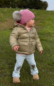Toddler on a grassy hillside wearing a pink SABBOT Frankie children's bobble hat with grey faux fur pom-poms