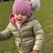 Laughing toddler running across the grass in a pink SABBOT Frankie children's double pom-pom bobble hat