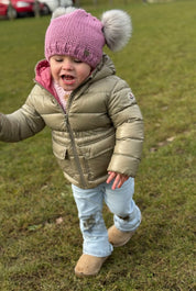 Laughing toddler running across the grass in a pink SABBOT Frankie children's double pom-pom bobble hat