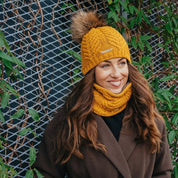 A smiling woman wearing the SABBOT Emma Bobble Hat in Mustard with the matching Emma Snood