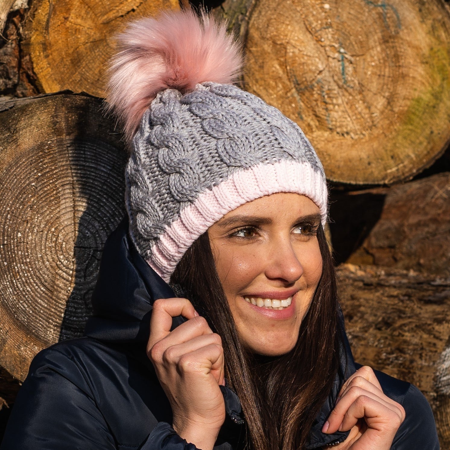 Woman smiling wearing the SABBOT Andrea chunky knit bobble hat in grey and pink with pink faux fur pom pom