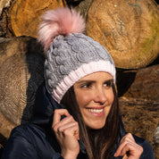 Woman smiling wearing the SABBOT Andrea chunky knit bobble hat in grey and pink with pink faux fur pom pom