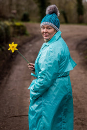 A woman holding a daffodil on a muddy track wearing the Andrea Bobble Hat in Teal/Grey