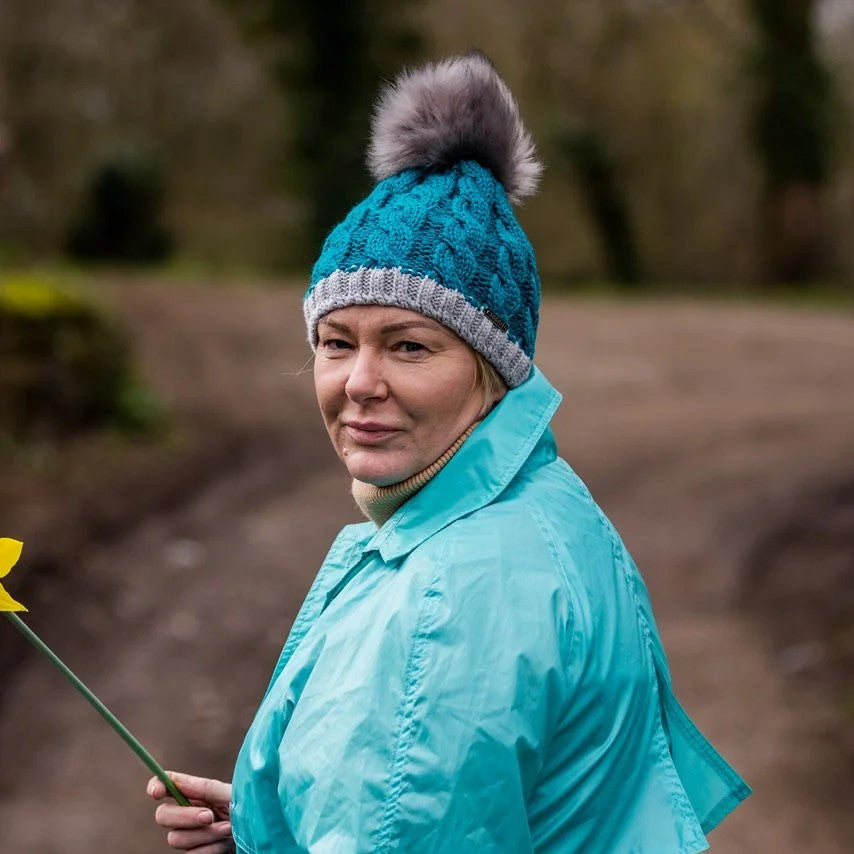 A woman holding a daffodil on a muddy track wearing the Andrea Bobble Hat in Teal/Grey