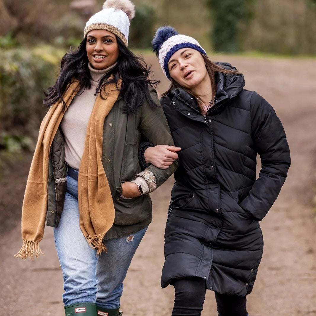 Two friends walking arm in arm down a country lane wearing the Andrea Bobble Hats in White/Camel and Navy/White