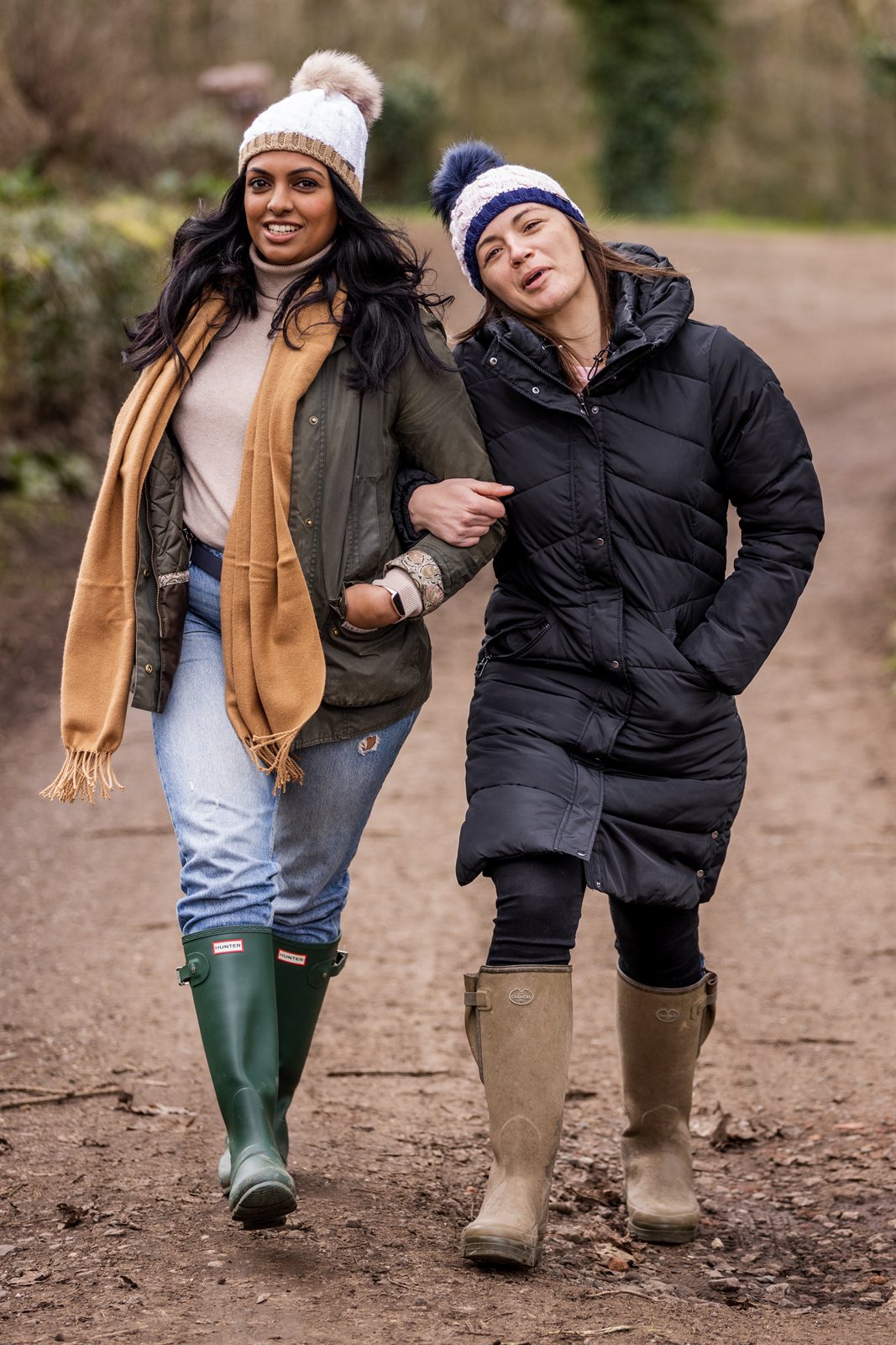 Two friends walking arm in arm down a country lane wearing the Andrea Bobble Hats in White/Camel and Navy/White