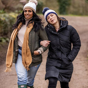 Two friends walking arm in arm down a country lane wearing the Andrea Bobble Hats in White/Camel and Navy/White