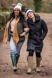 Two friends walking arm in arm down a country lane wearing the Andrea Bobble Hats in White/Camel and Navy/White