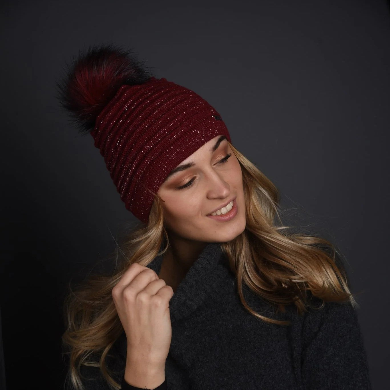 A smiling woman wearing the SABBOT Blanka sparkle knit bobble hat in Burgundy