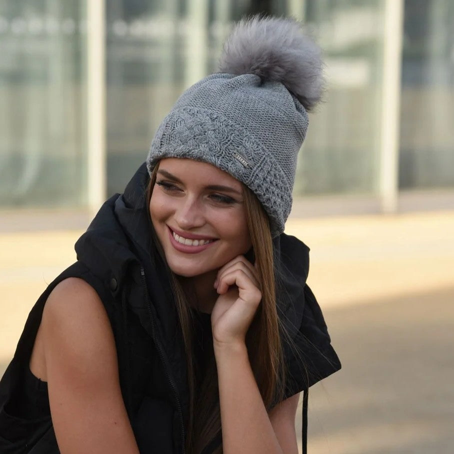Woman smiling outdoors wearing the SABBOT Lenka knit bobble hat with faux fur pom pom