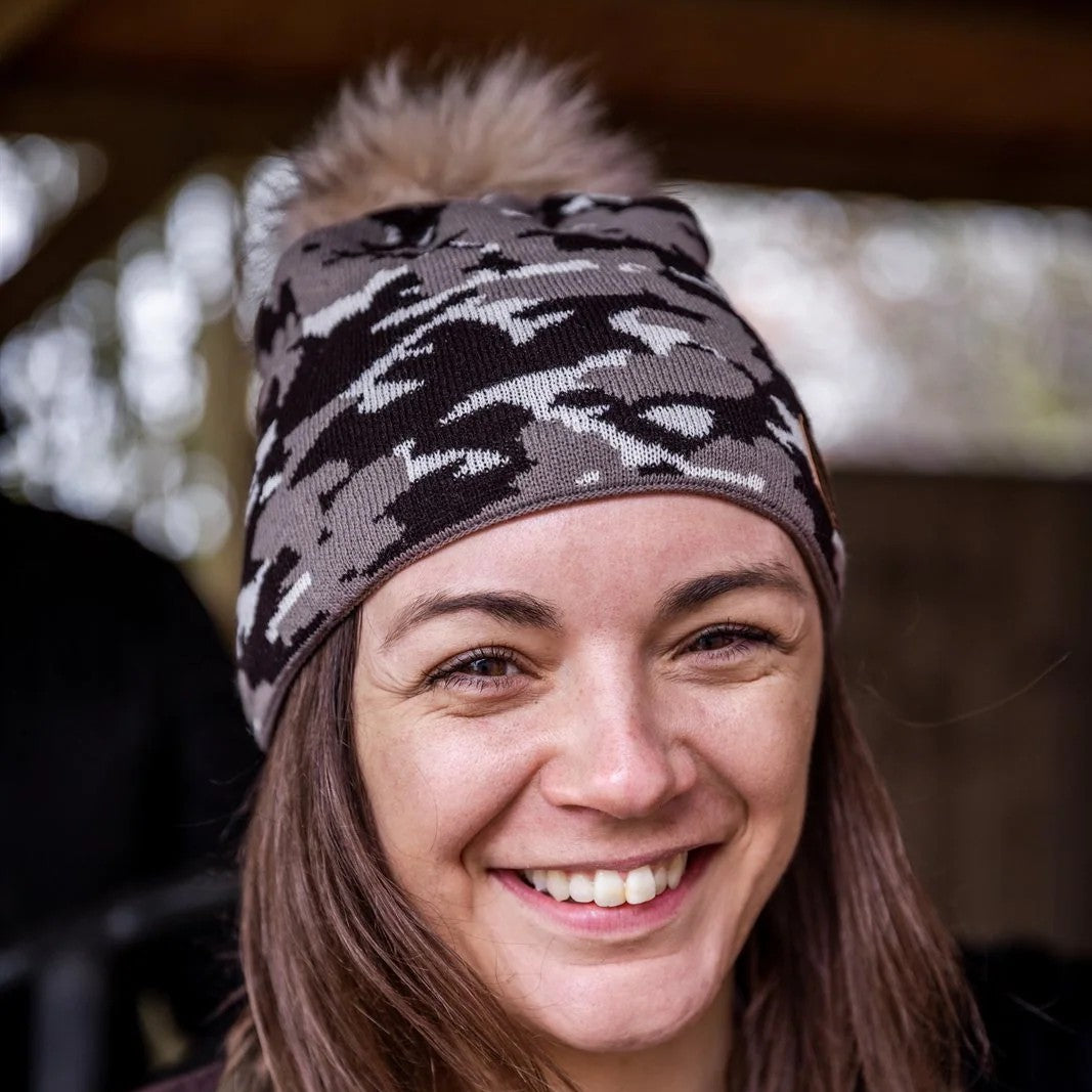 A smiling woman under a barn roof wearing the Calli Bobble Hat in Brown Camo