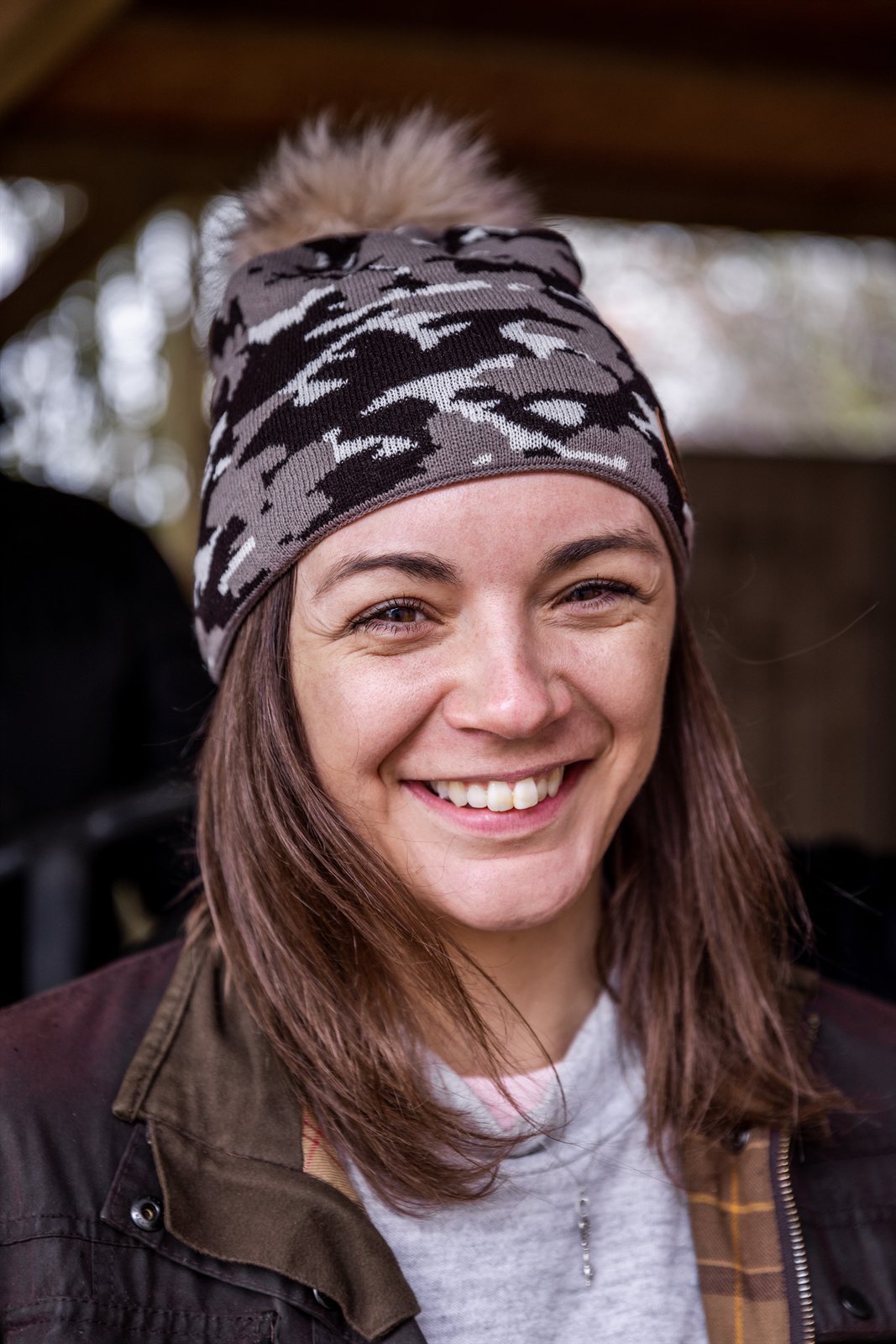 A smiling woman under a barn roof wearing the Calli Bobble Hat in Brown Camo