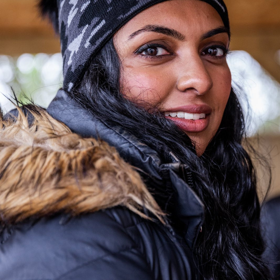A smiling woman under a barn roof wearing the Calli Bobble Hat in Grey Camo