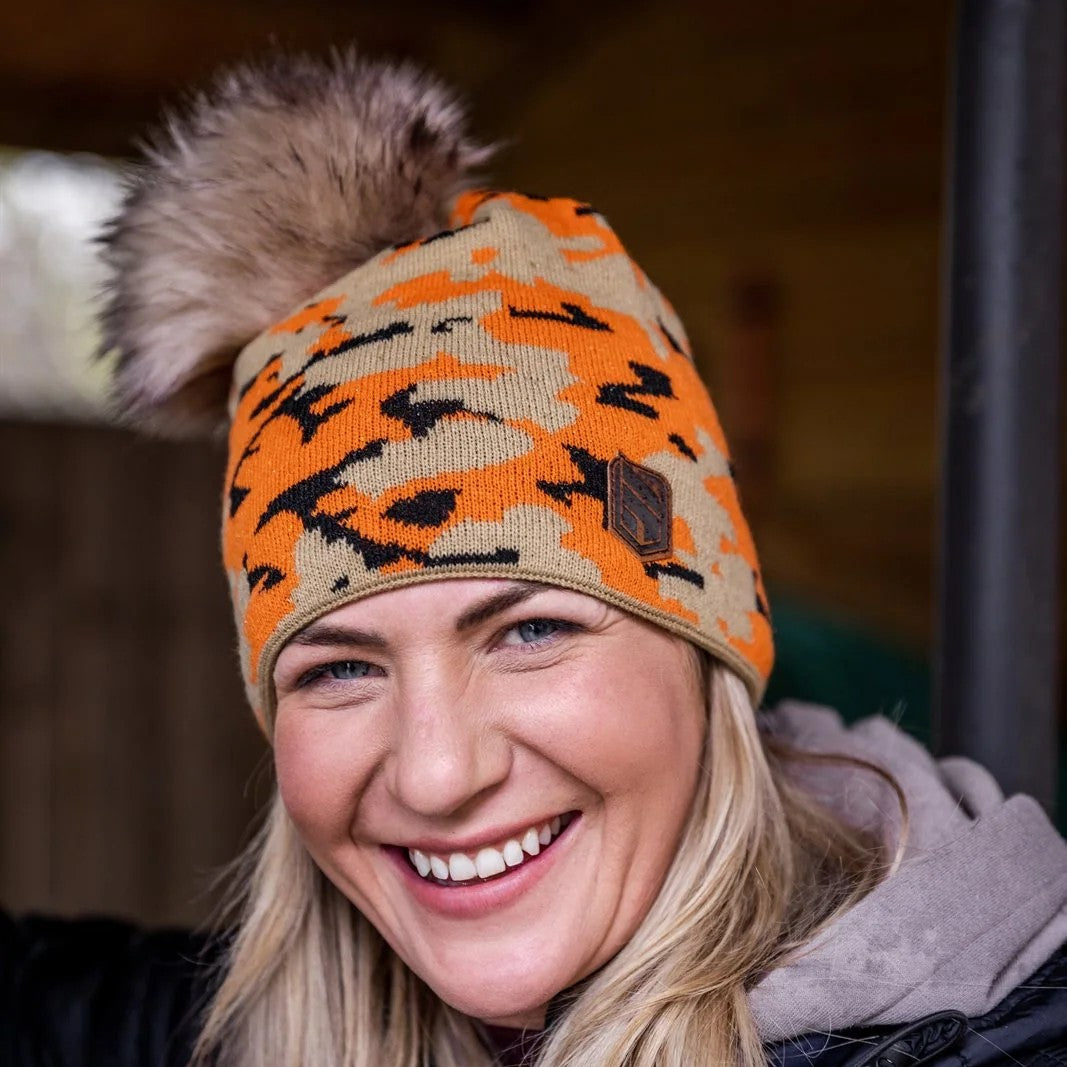 A smiling woman under a barn roof wearing the Calli Bobble Hat in Orange Camo