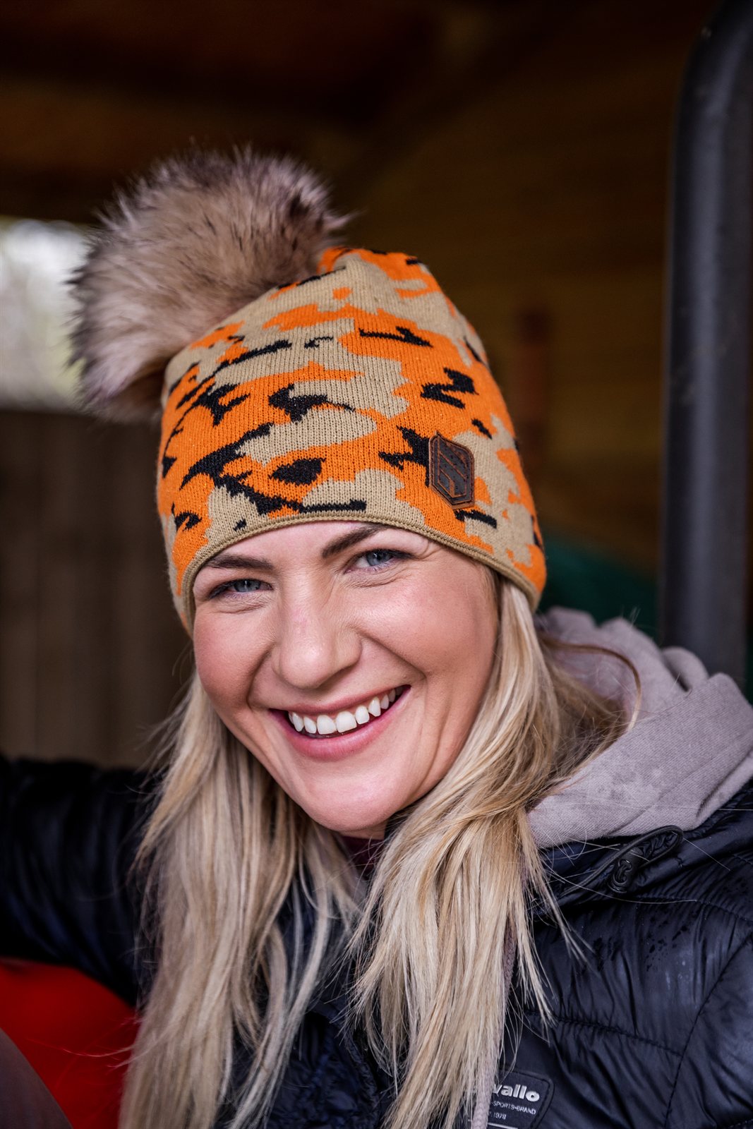 A smiling woman under a barn roof wearing the Calli Bobble Hat in Orange Camo
