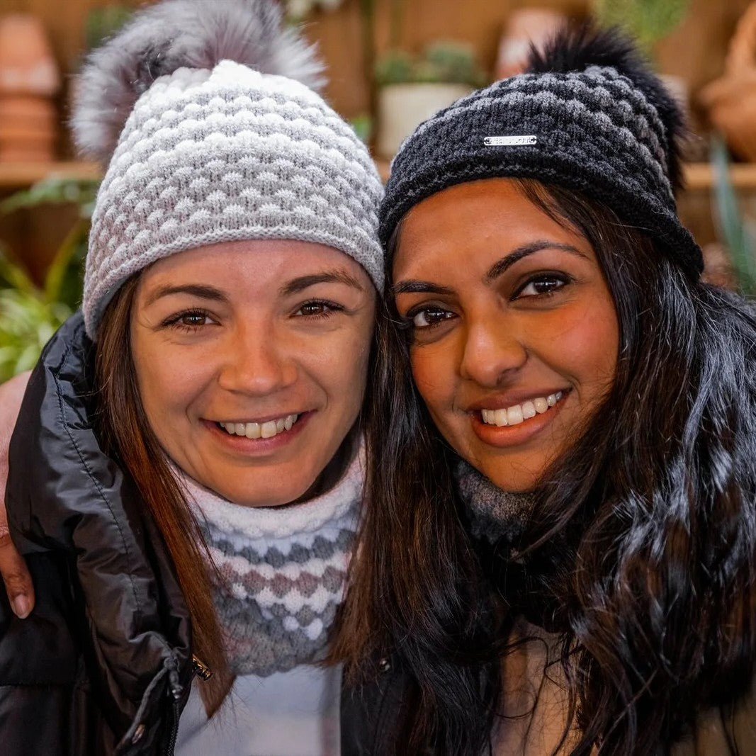 Two friends laughing in a plant-filled glasshouse wearing the Chloe Bobble Hats in Grey/White and Black/Grey