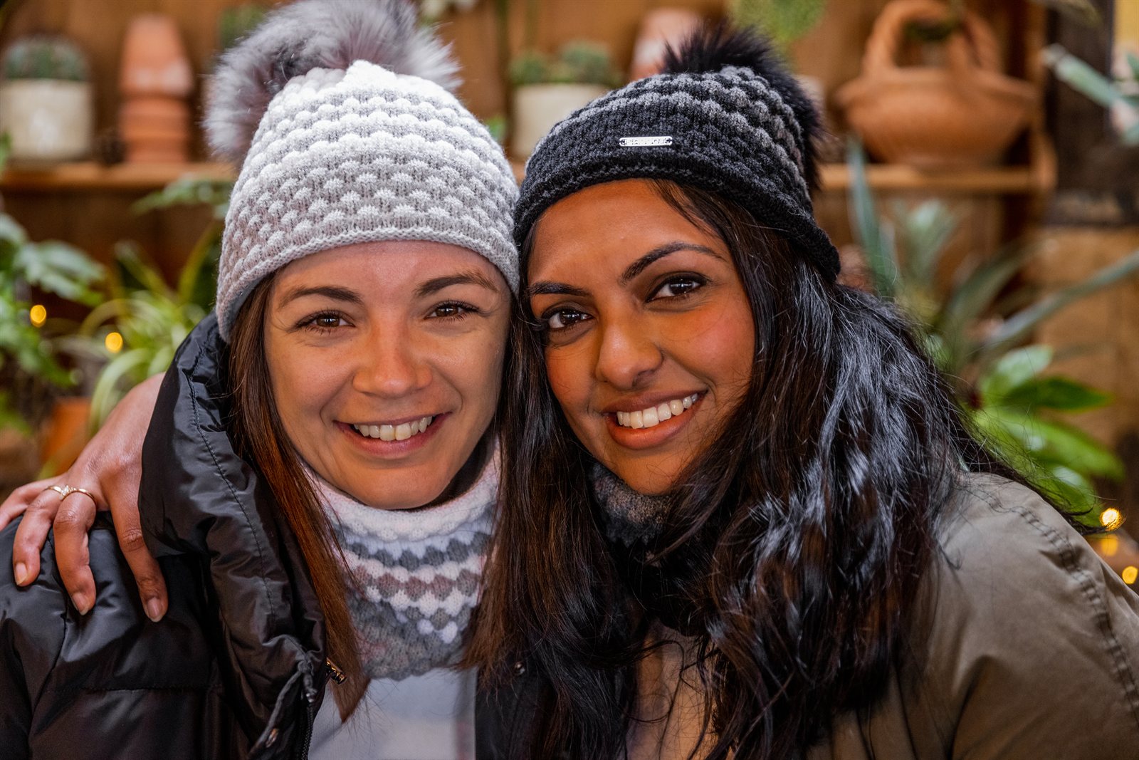 Two friends laughing in a plant-filled glasshouse wearing the Chloe Bobble Hats in Grey/White and Black/Grey