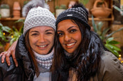 Two friends laughing in a plant-filled glasshouse wearing the Chloe Bobble Hats in Grey/White and Black/Grey