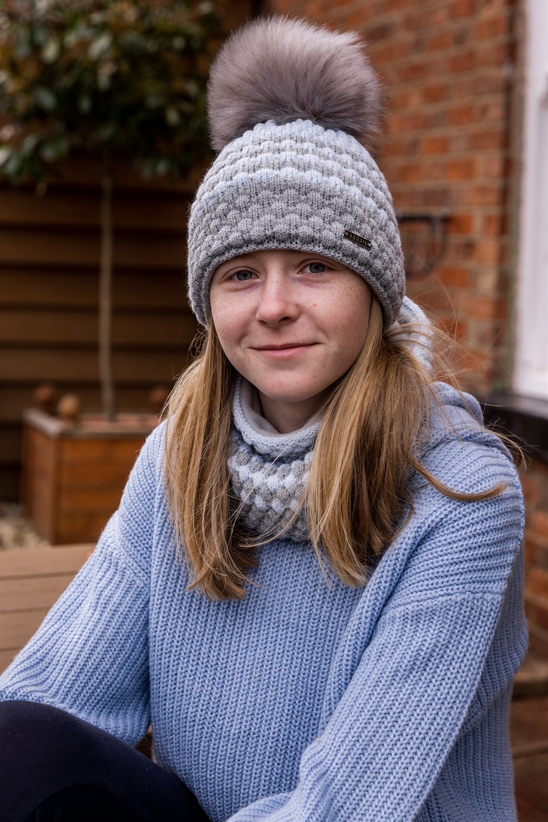 A teenager sitting on a garden bench wearing the Chloe Bobble Hat and Snood in Grey/White