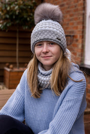 A teenager sitting on a garden bench wearing the Chloe Bobble Hat and Snood in Grey/White