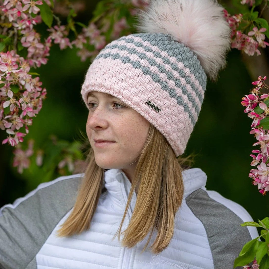Woman among spring blossom wearing the SABBOT Chloe bobble hat in pink and grey
