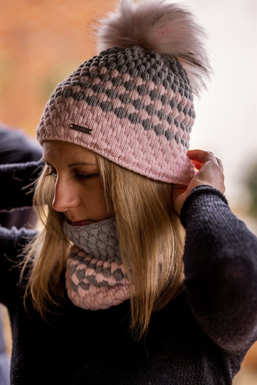 A woman adjusting her pink and grey bubble-knit hat wearing the Chloe Bobble Hat and Snood in Pink/Grey