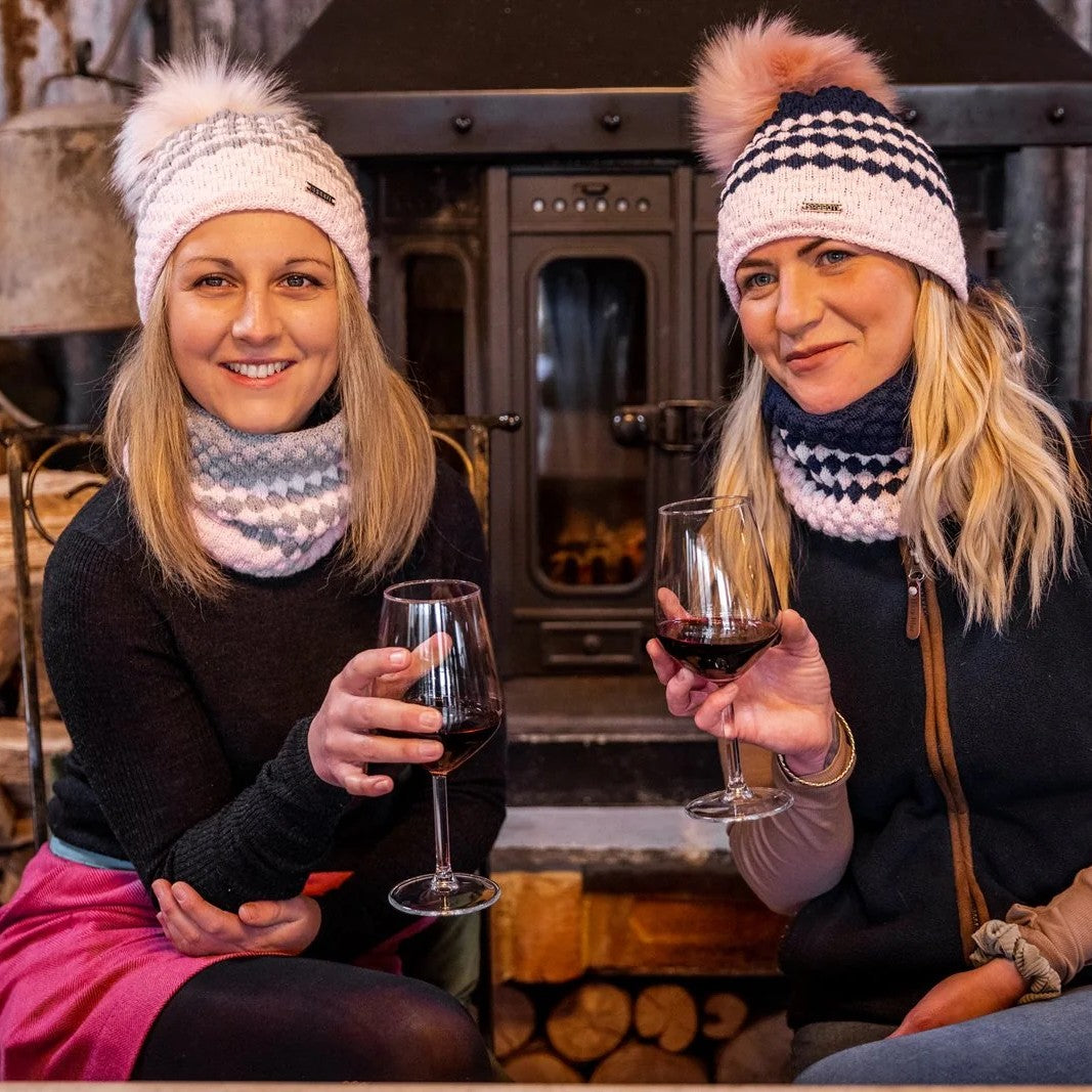 Two friends with wine beside a wood stove wearing SABBOT Chloe Bobble Hats