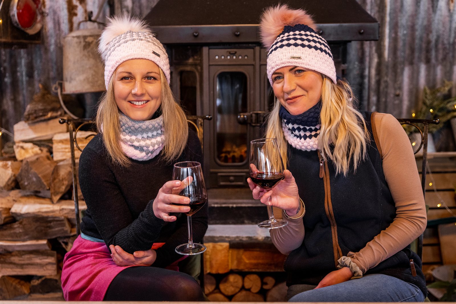 Two friends with wine beside a wood stove wearing SABBOT Chloe Bobble Hats
