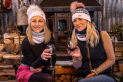 Two friends with wine beside a wood stove wearing SABBOT Chloe Bobble Hats