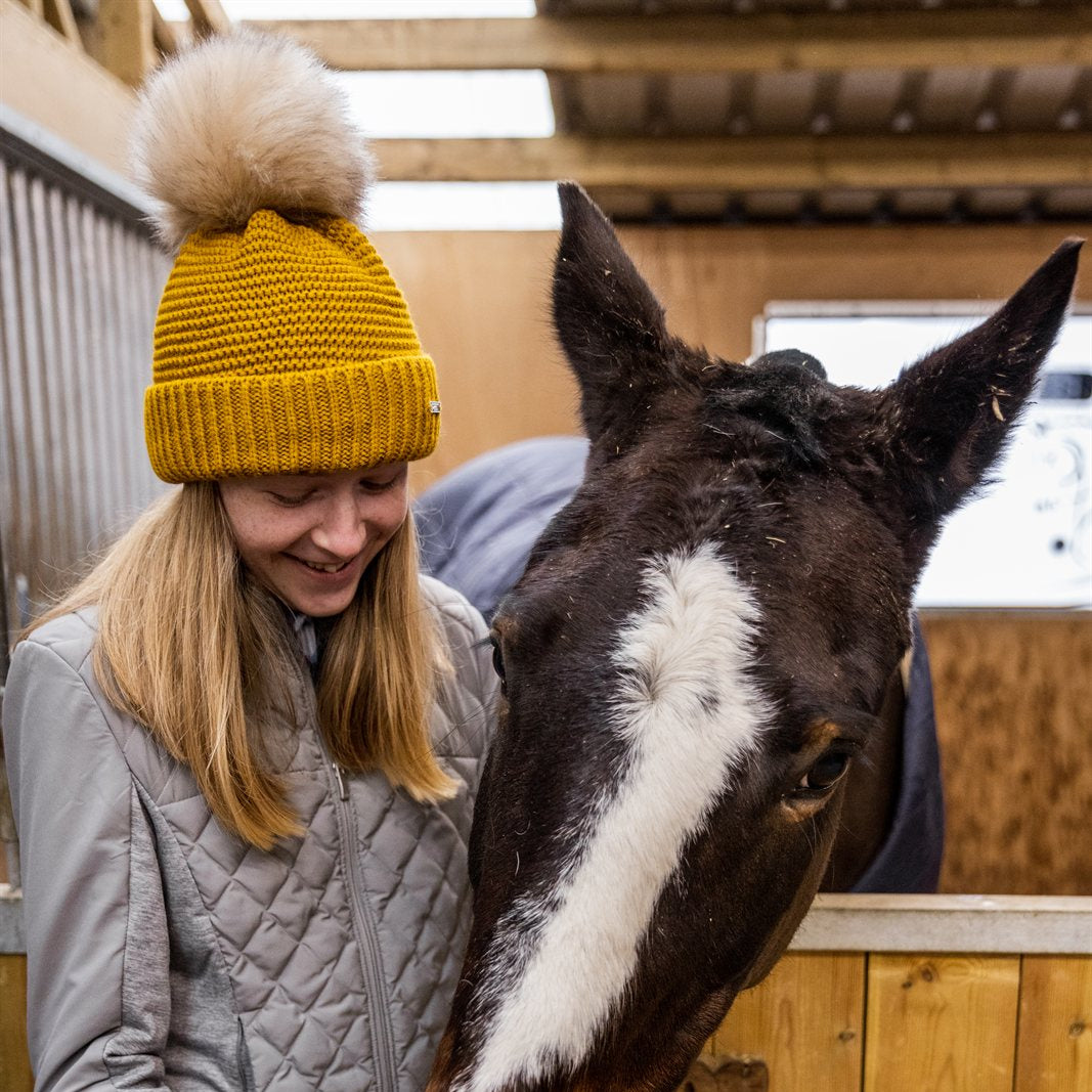 A woman with a horse in the stables wearing the Eva Bobble Hat in Mustard