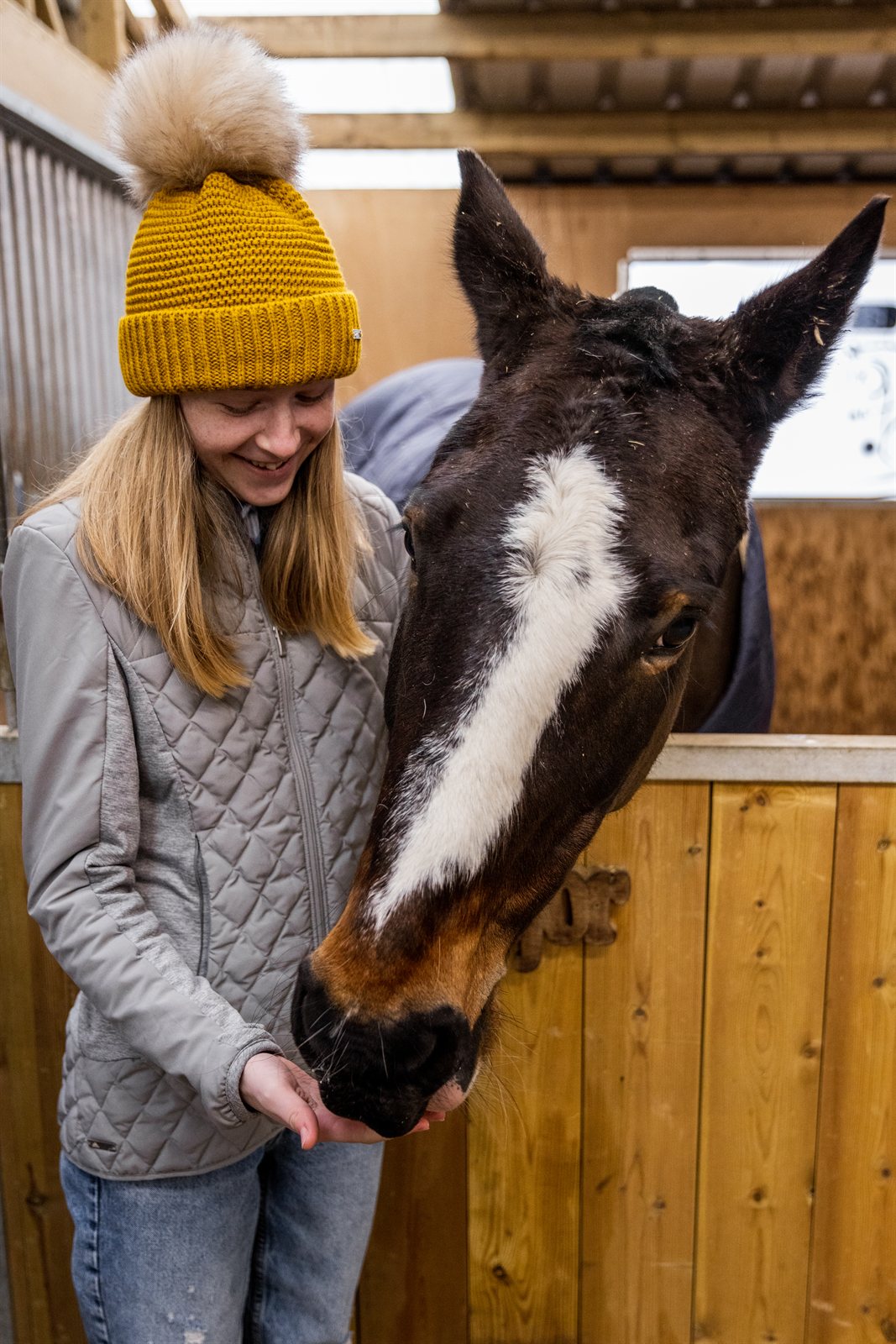 A woman with a horse in the stables wearing the Eva Bobble Hat in Mustard