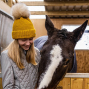 A woman with a horse in the stables wearing the Eva Bobble Hat in Mustard