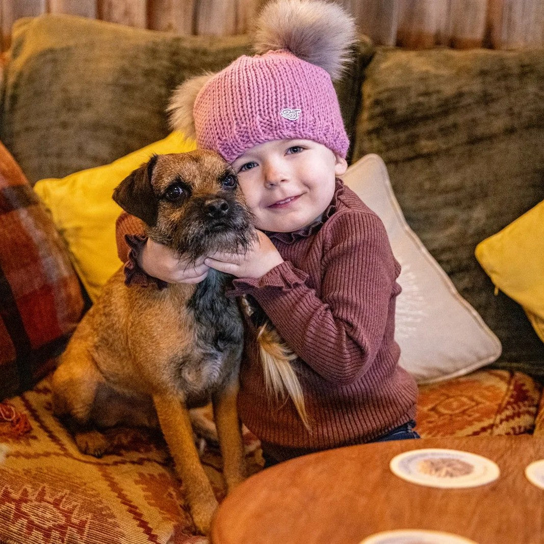 A child cuddling a terrier on a cushioned sofa wearing the Frankie Childrens Bobble Hat in Pink