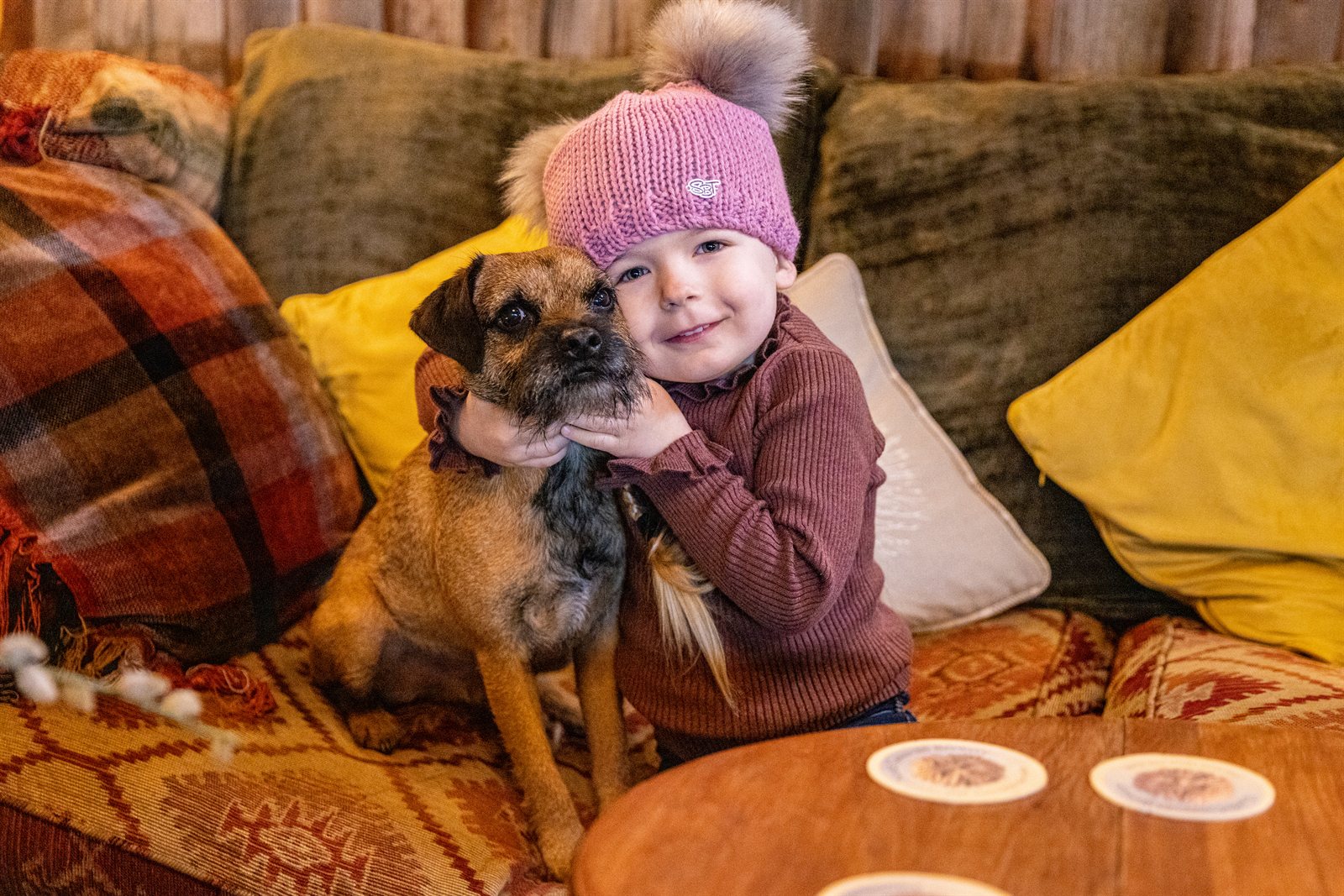 A child cuddling a terrier on a cushioned sofa wearing the Frankie Childrens Bobble Hat in Pink