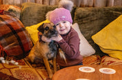 A child cuddling a terrier on a cushioned sofa wearing the Frankie Childrens Bobble Hat in Pink