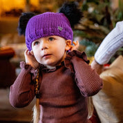 A toddler pulling on a purple double pom-pom hat wearing the Frankie Childrens Bobble Hat in Purple