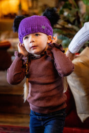 A toddler pulling on a purple double pom-pom hat wearing the Frankie Childrens Bobble Hat in Purple