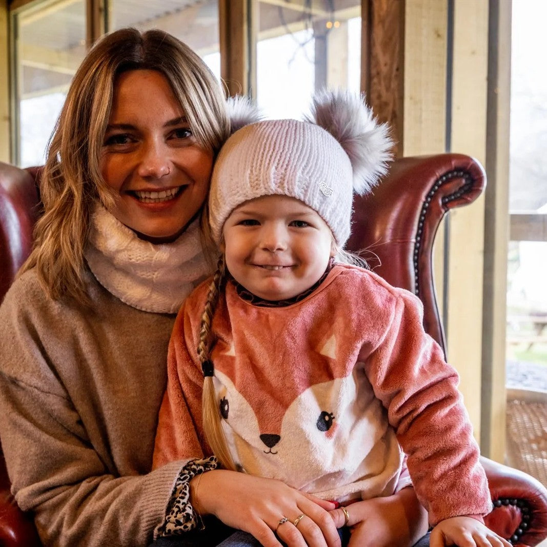 A toddler in a white double pom-pom hat with her mum wearing the Frankie Childrens Bobble Hat in White