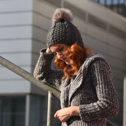Woman in a long knitted coat wearing the SABBOT Frida bobble hat in dark grey