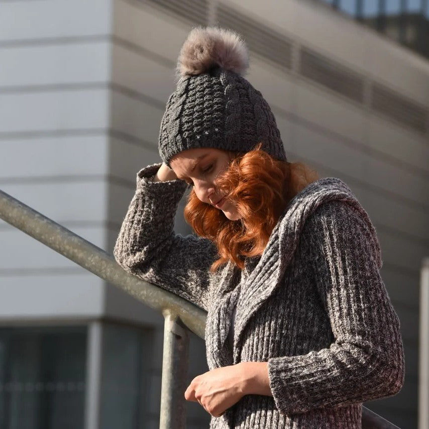 Woman in a long knitted coat wearing the SABBOT Frida bobble hat in dark grey